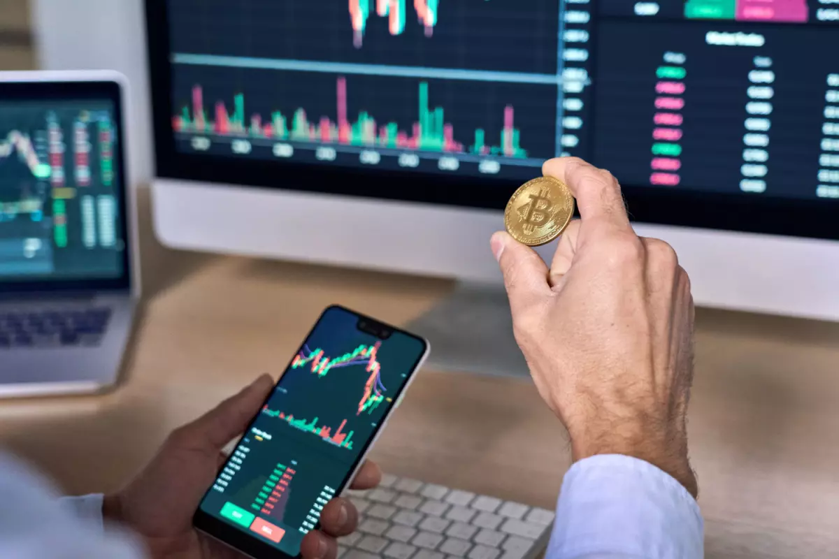 Crypto investor holding Bitcoin and cellphone with stock market on it